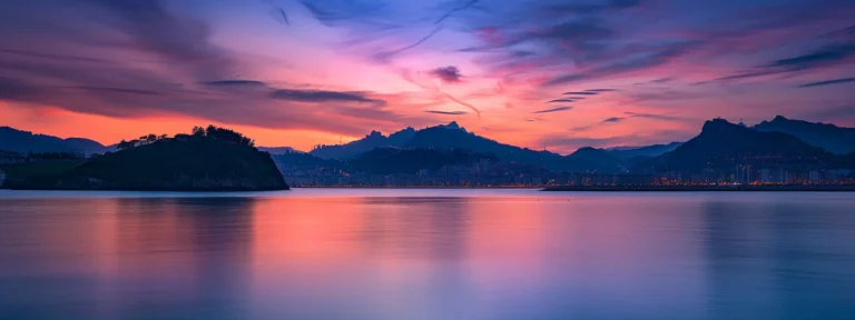 un vibrante atardecer sobre la bahía de san sebastián, donde las majestuosas montañas se reflejan en las aguas tranquilas, enmarcando una hermosa vista de la ciudad y sus icónicas atracciones turísticas.