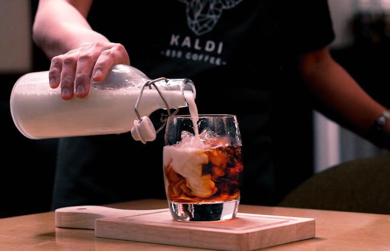 A barista skillfully pours milk into a glass of iced coffee on a wooden board.